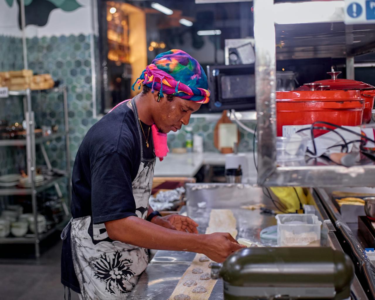 services-06 A chef wears colorful headscarf and apron while crafting pastries in a professional kitchen.