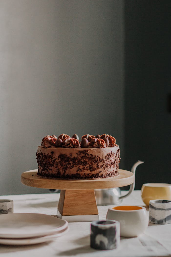 Tasty cake decorated with chocolate frosting served on wooden stand among ceramic plates and teapot for teatime