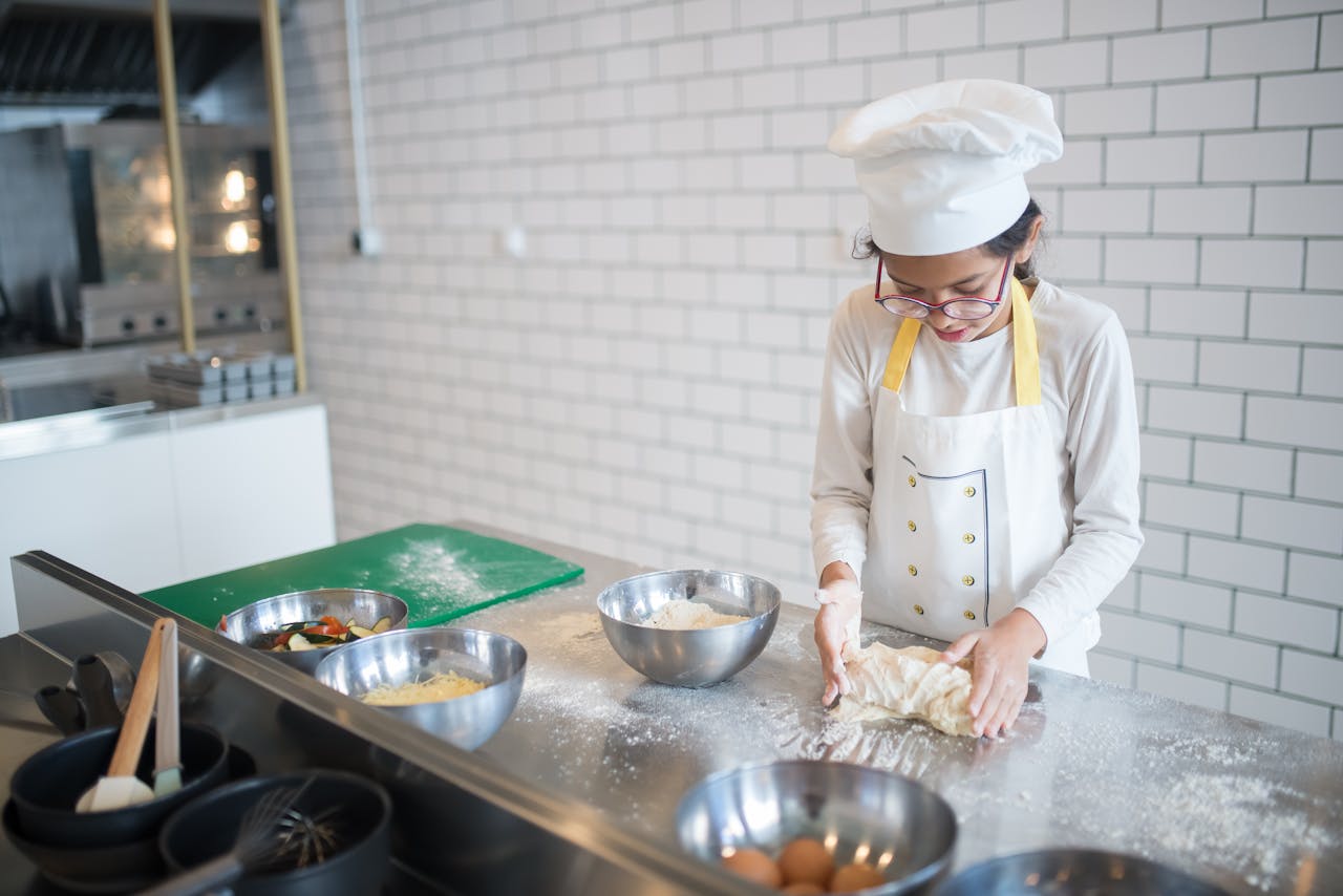 about-us A young girl in a chef's hat kneads dough in a contemporary kitchen setting.